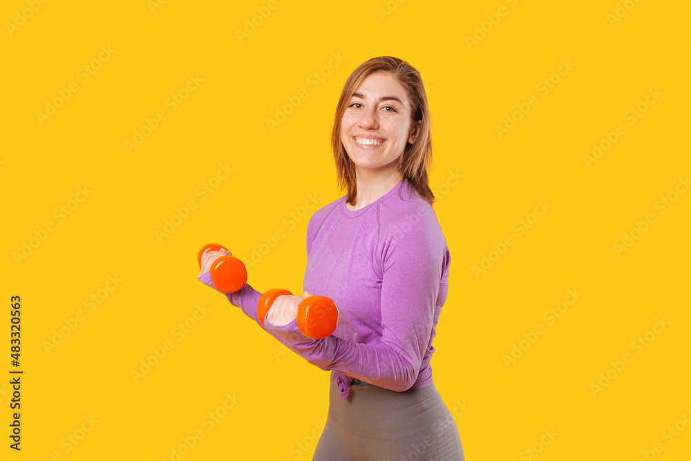 Young smiling woman is lifting up some red dumbbells while smiling at the camera. Portrait over yellow background. Young smiling woman is lifting up some red dumbbells while smiling at the camera. Portrait over yellow background.
