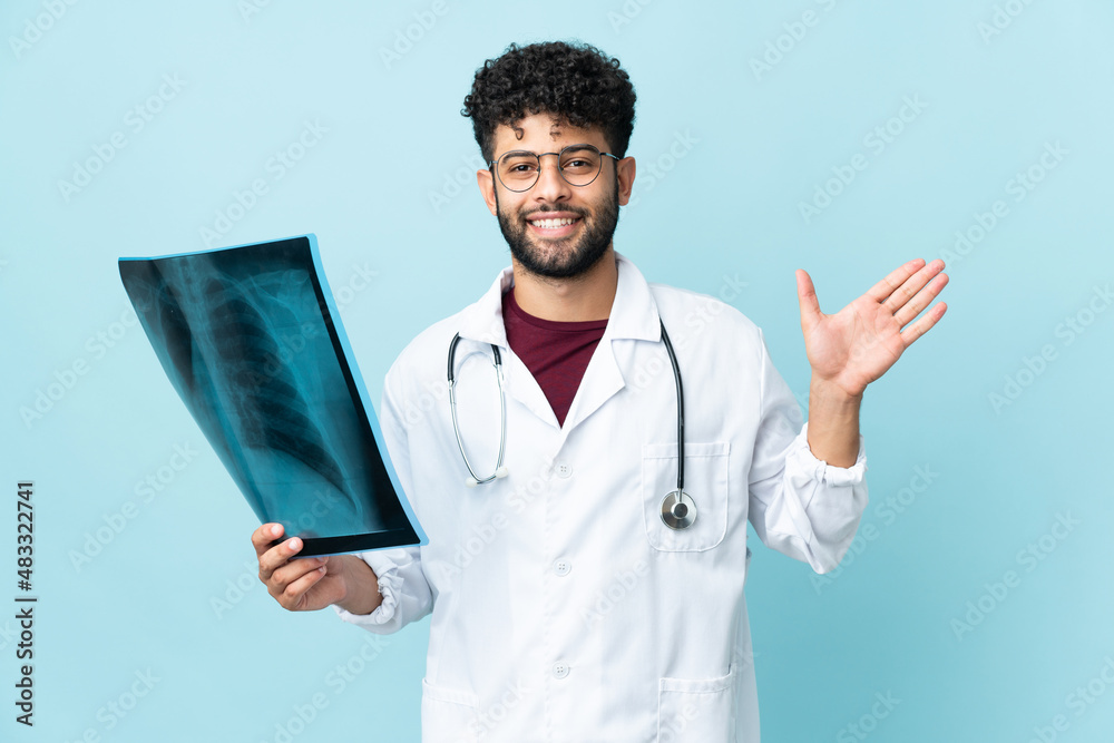 Young Moroccan traumatologist in workplace isolated on blue background saluting with hand with happy expression Young Moroccan traumatologist in workplace isolated on blue background saluting with hand with happy expression