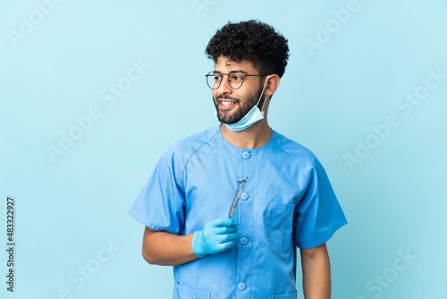 Moroccan dentist man holding tools isolated on blue background looking to the side and smiling