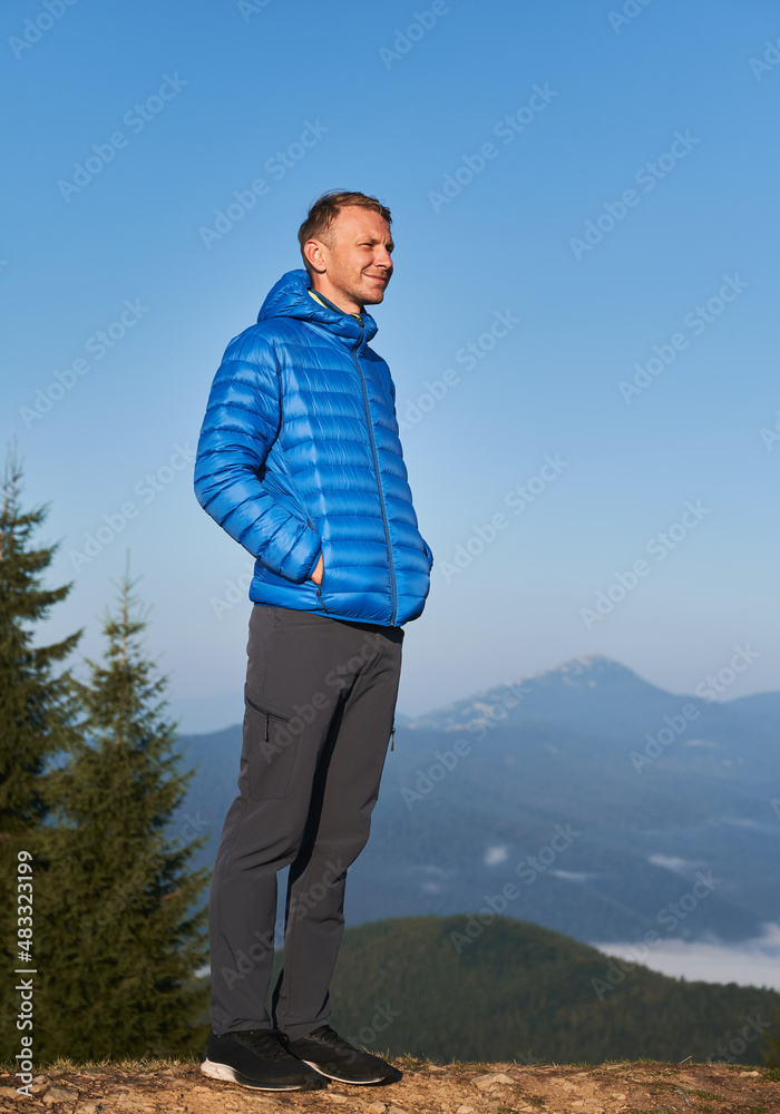 Young man looking to the right with smile, squinting from bright rays of sun against the backdrop of mountain peaks and cloudless blue sky. Walking in the autumn mountains. Young man looking to the right with smile, squinting from bright rays of sun against the backdrop of mountain peaks and cloudless blue sky. Walking in the autumn mountains.