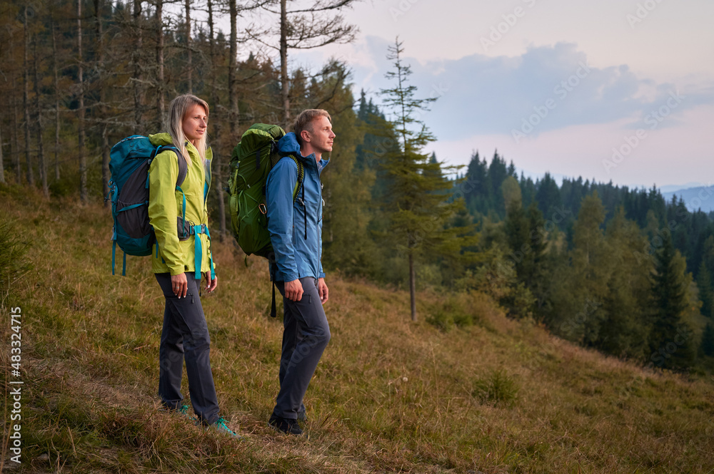 Full length of young couple travelers standing on grassy hill and enjoying beauty of nature. Young woman and man with backpacks hiking together in mountains. Concept of travelling and active leisure. Full length of young couple travelers standing on grassy hill and enjoying beauty of nature. Young woman and man with backpacks hiking together in mountains. Concept of travelling and active leisure.