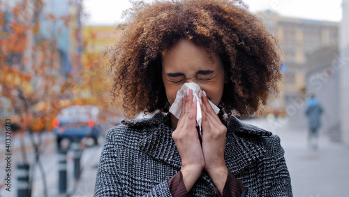 Sick ill african american woman with curly hair blowing runny nose into tissue paper outdoors cold allergies respiratory virus girl suffers from contagious rhinitis symptom autumn in city flu pandemic photo