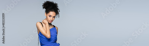 curly african american model in bright blue dress looking at camera while posing isolated on grey, banner.