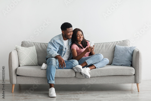 African american couple sitting on couch, sharing using cellphone