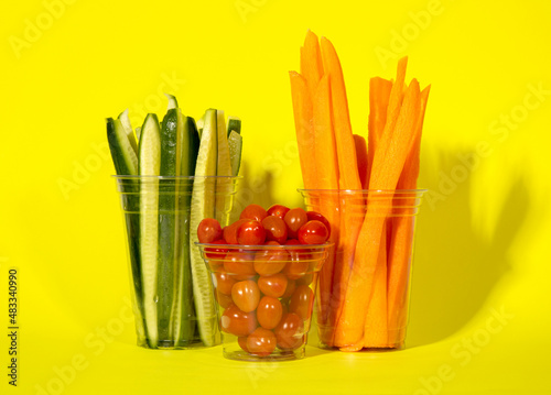 Tomatoes, cucumbers, carrots in plastic cups. Plastic cup with background. Plastic cup and vegetables. Snack serving containers. Full plastic cup on colored background. Food in a clear glass