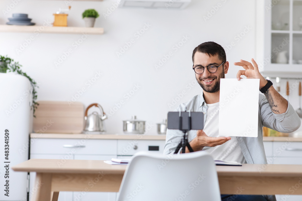 Smiling mature caucasian male teacher with beard in glasses showing blank paper to smartphone webcam Smiling mature caucasian male teacher with beard in glasses showing blank paper to smartphone webcam