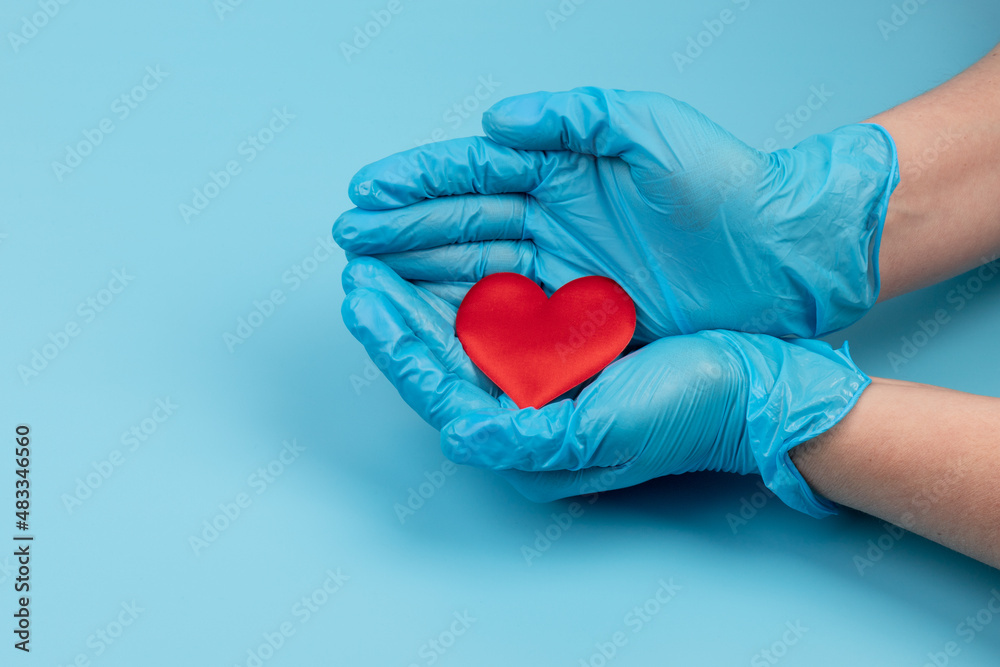 Fototapeta premium Red heart in hands in blue medical gloves on blue background. Background for the day of the medic. World Heart Day. Heart diseases.