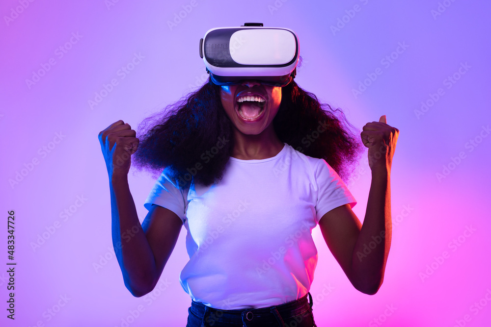 Young black lady in VR glasses exploring cyberspace, shouting in excitement, playing virtual reality game in neon light