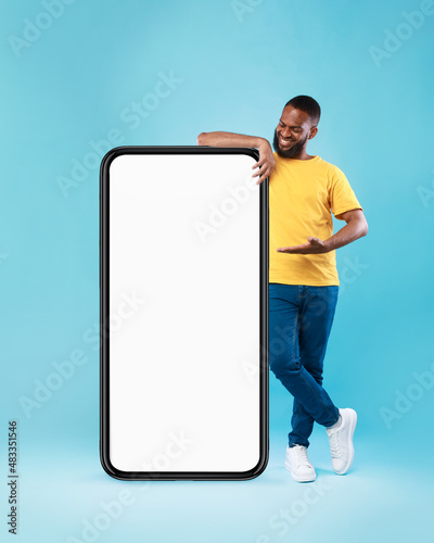 Black guy presenting giant smartphone with empty screen on blue background, mockup for online ad, website or mobile app