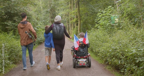 A family with a disabled person moves through the forest in the park. They take steps, the carriage rolls. Leisure activities in the city park. Family communication.