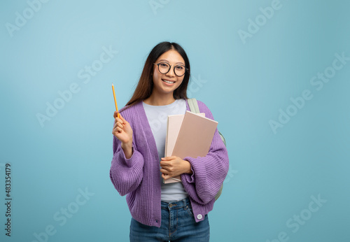 I know the answer. Excited asian female student in eyeglasses holding notepad and pointing pencil up, blue background
