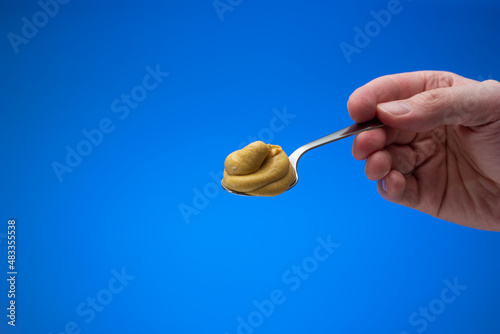 Mustard paste on a small silver spoon held in hand by man. Close up studio shot, isolated on blue background