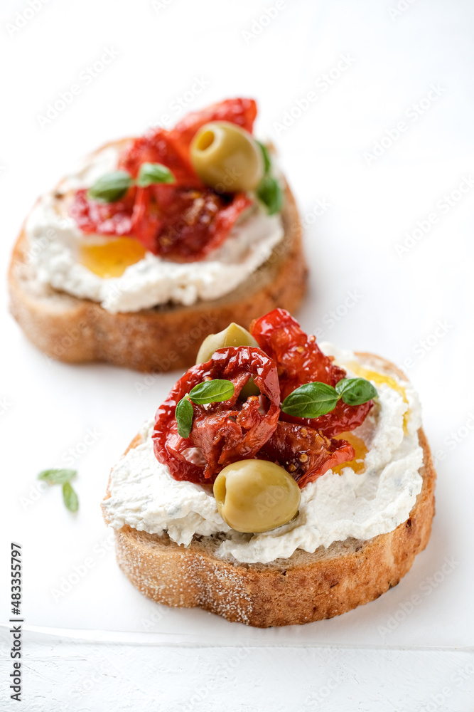 Two bruschettas with ricotta cheese, olive and dried tomatoes on ciabatta bread on white background. Olive oil and spices decorated. Copyspace.