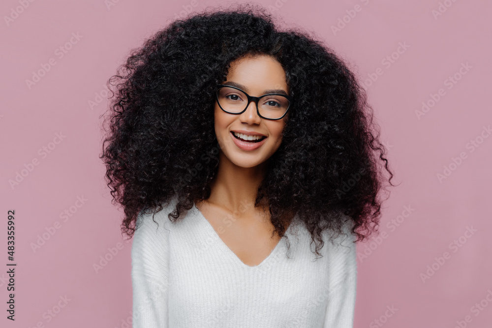 Portrait of pleasant looking smiling woman with Afro hairstyle, wears optical glasses and white sweater, has cheerful face expression, isolated over purple background. Millennial girl poses indoor Portrait of pleasant looking smiling woman with Afro hairstyle, wears optical glasses and white sweater, has cheerful face expression, isolated over purple background. Millennial girl poses indoor