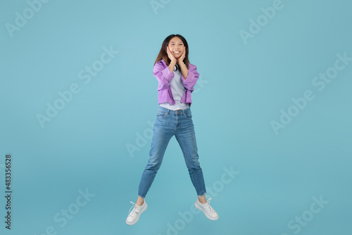 Shocking sale. Surprised amazed young asian lady jumping in air and touching face with amazement, blue background