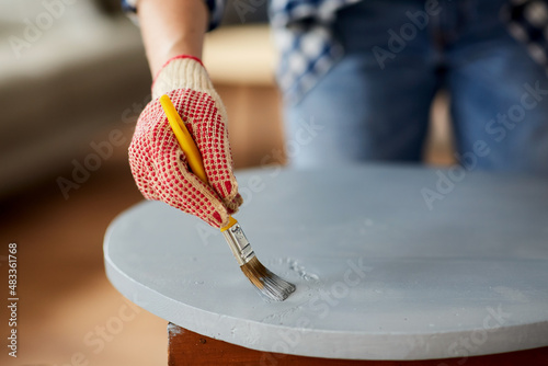 furniture renovation, diy and home improvement concept - close up of woman in gloves with paint brush painting old wooden table in grey color