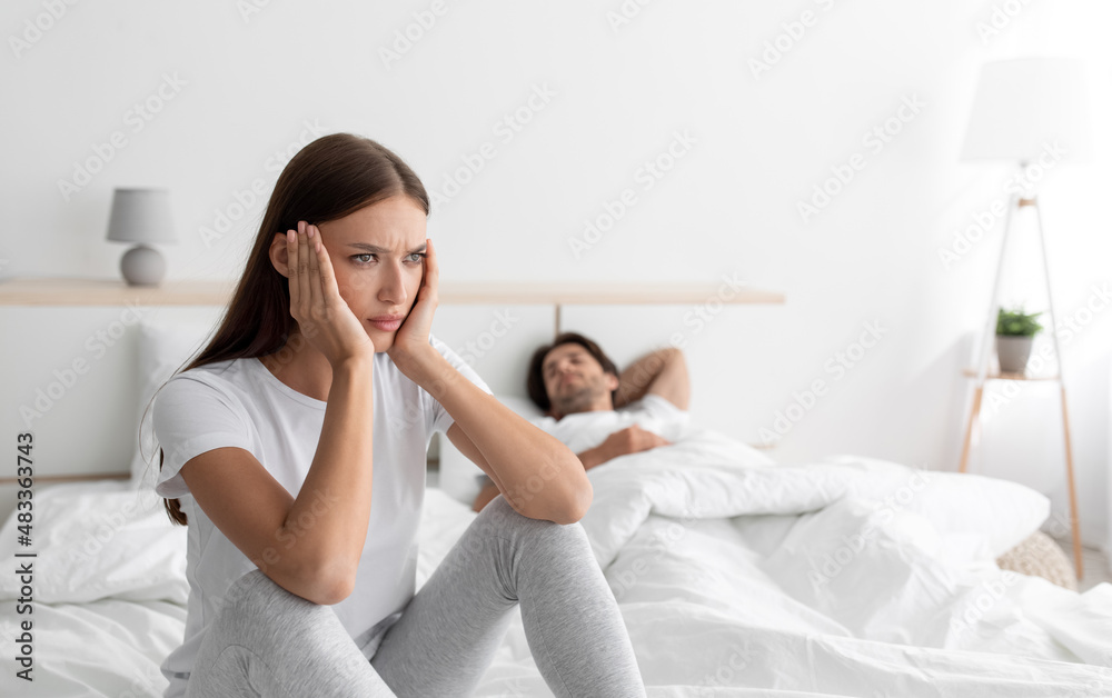 Sad offended european lady sitting on bed with sleeping husband in bedroom interior, free space Sad offended european lady sitting on bed with sleeping husband in bedroom interior, free space