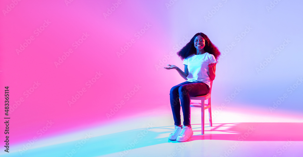 Happy black lady sitting on chair and offering empty space for your advertisement in neon light, banner design