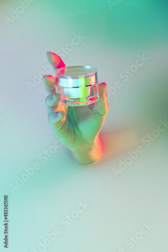 Close up female hand holding cosmetic jar appears from milk bath with soft white glow in neon light. Beaty, cosmetics, fashion, art