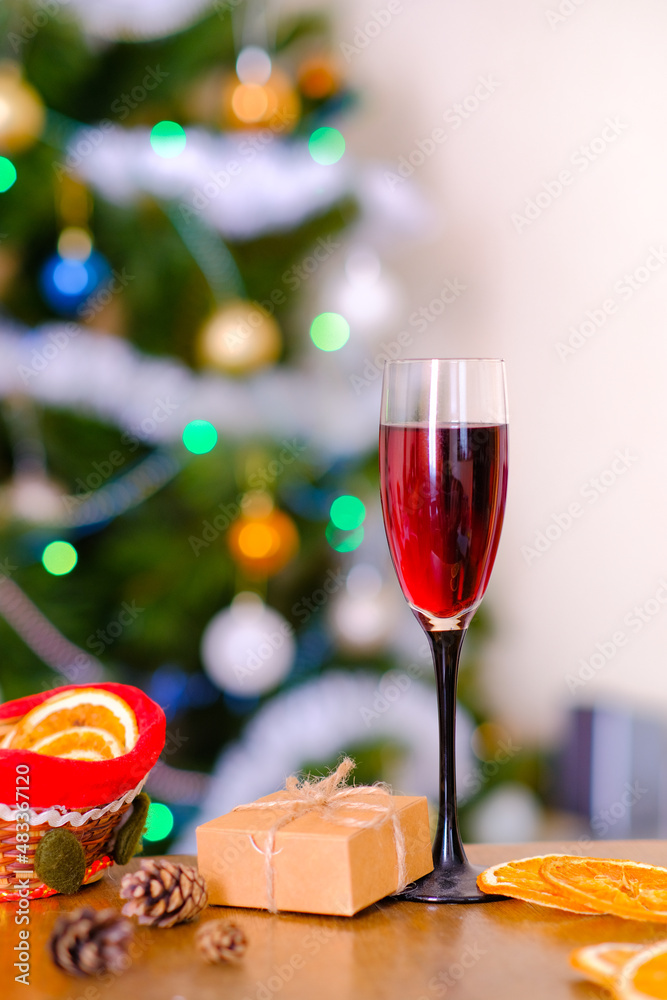 A glass with red wine stand on a wooden table against the background of a festive elegantly decorated Christmas tree. Blur. Bokeh effect. Small beige neutral minimalistic gift, cones, dried oranges.