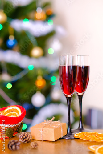 Two glasses with red wine stand on a wooden table against the background of a Christmas tree. Festive elegantly decorated Christmas tree. Blur. Bokeh effect. Small beige neutral minimalistic gift