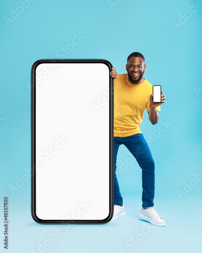 Handsome black guy standing near giant cellphone with blank screen, showing mobile device on blue background, mockup