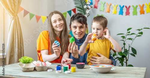 Mom  dad and little son in bright clothes is coloring eggs in a decorated room. Concept of family preparation for Easter  festive spring mood