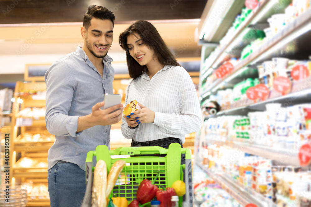 Arab Couple Using Smartphone Buying Food Shopping In Modern Supermarket