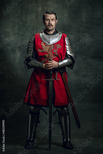 Portrait of brutal seriuos man, medieval warrior or knight with dirty wounded face holding sword isolated over dark background. Comparison of eras