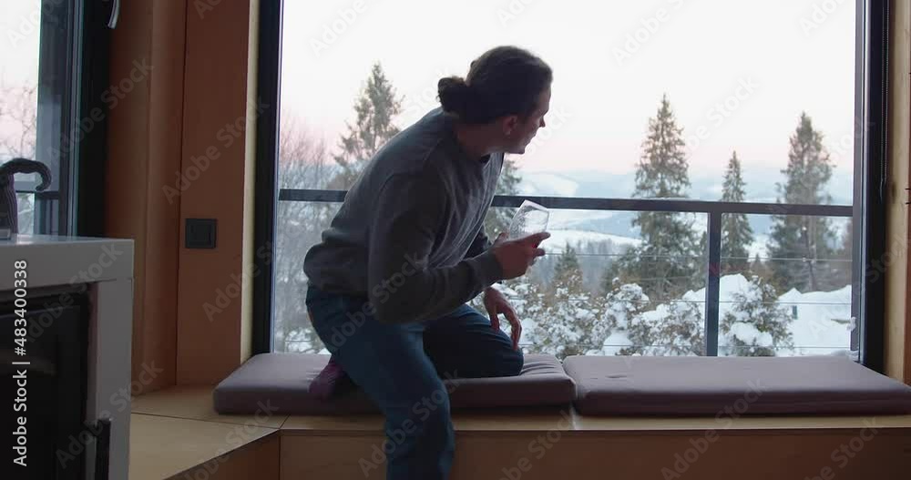 man with tied hair in a gray sweater and jeans sits on a soft window sill in a house with a glass of red wine against the backdrop of winter mountains and forest. overall plan man with tied hair in a gray sweater and jeans sits on a soft window sill in a house with a glass of red wine against the backdrop of winter mountains and forest. overall plan