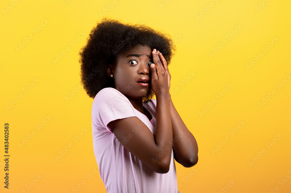 Stressed young black lady covering face with hands and peeking with one eye, standing on yellow background Stressed young black lady covering face with hands and peeking with one eye, standing on yellow background