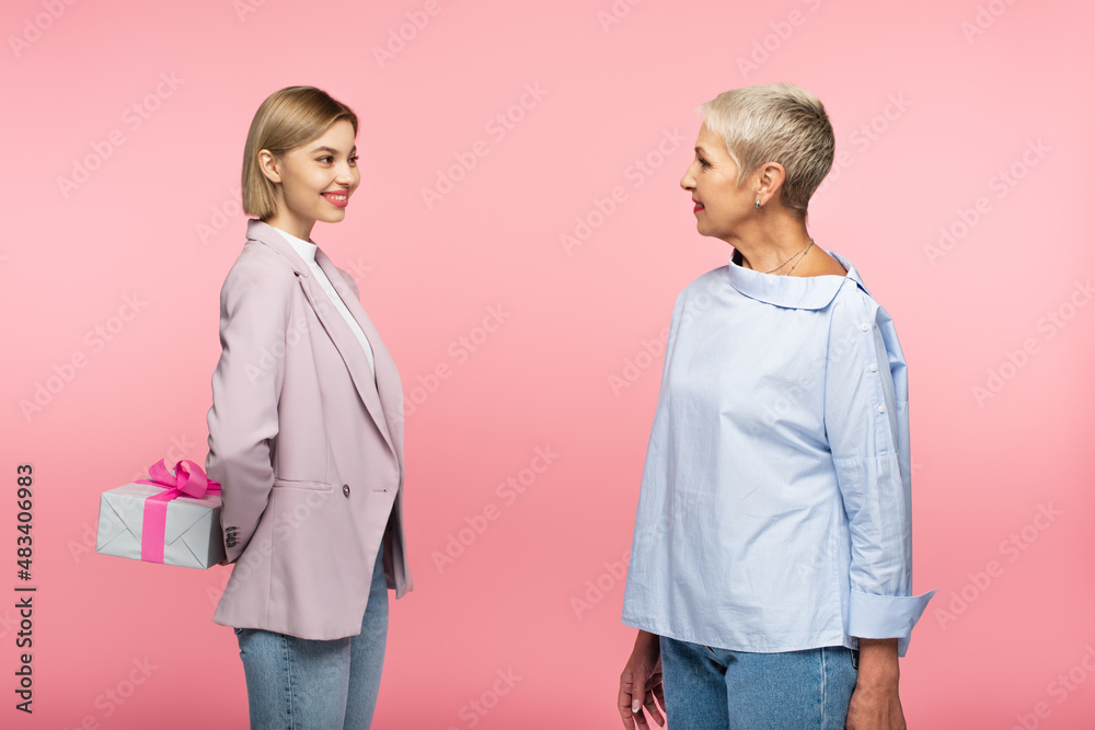 cheerful young woman holding present behind back near mature mother isolated on pink cheerful young woman holding present behind back near mature mother isolated on pink