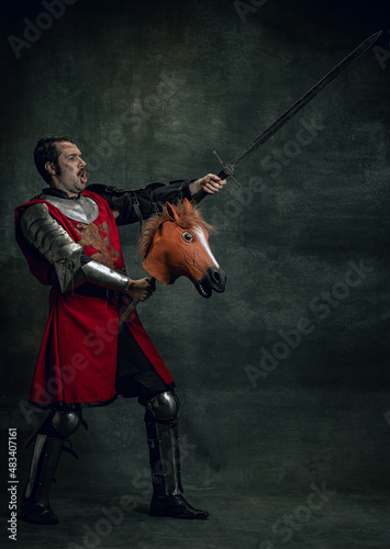 Portrait of medieval warrior or knight wearing helmet and armor riding toy horse, holding big sword isolated over dark background. Comparison of eras, history © master1305