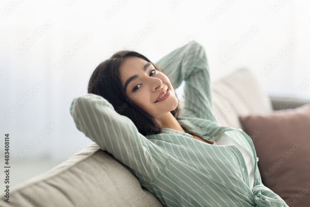 Lazy weekend at home. Charming Indian woman resting with hands behind head on cozy couch at living room Lazy weekend at home. Charming Indian woman resting with hands behind head on cozy couch at living room