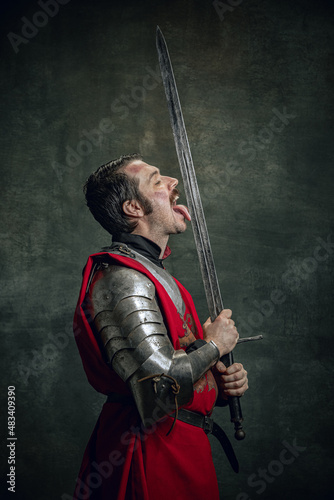 Comic portrait of funny medieval warrior or knight with dirty wounded face holding sword isolated over dark background. Comparison of eras