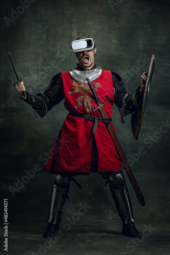 Portrait of man, medieval warrior, knight with dirty wounded face in VR headset isolated over dark vintage background. Comparison of eras, history