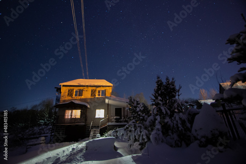 Night shot of a countryside brick house with lighted windows with a starry sky on a background in winter. Concept of peace, magic, idyllic, home, dreams.