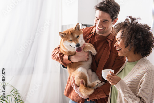 cheerful man holding funny shiba inu dog near african american woman with cup of coffee