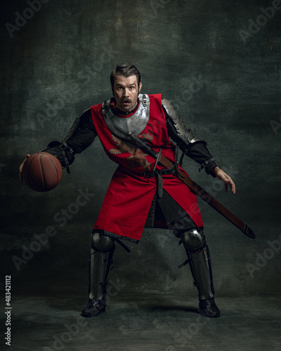 Portrait of medieval warrior or knight with dirty wounded face playing with basketball ball isolated over dark background. Comparison of eras, history photo