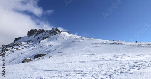 Mountaineers walking, hiking on a winter sunny day to reach the mountain peak. Wind and clouds are passing in the sky. Traveling and connecting with nature. 
