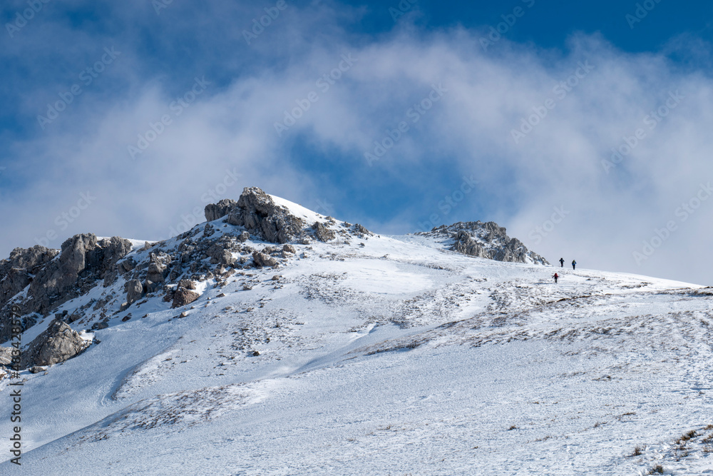 Mountaineers walking, hiking on a winter sunny day to reach the mountain peak. Wind and clouds are passing in the sky. Traveling and connecting with nature. 
