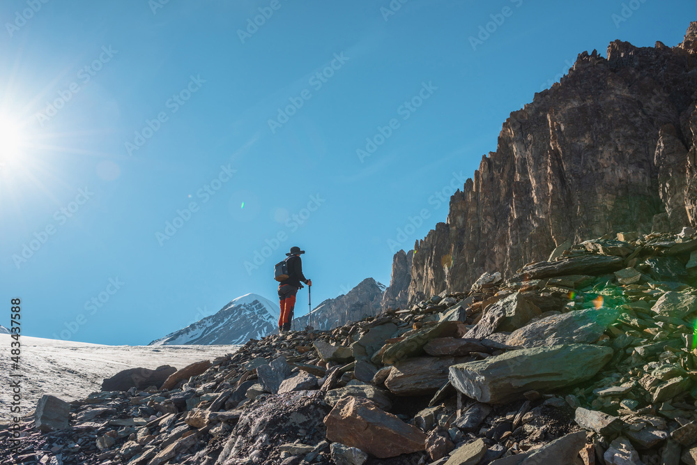 Scenic alpine landscape with silhouette of hiker with trekking poles against large sharp rocks and snow mountain peak in sunlight. Man with backpack in high mountains under blue sky in sunny day.