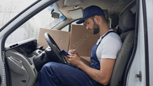 Side view of delivery man writing on clipboard near carton boxes in car