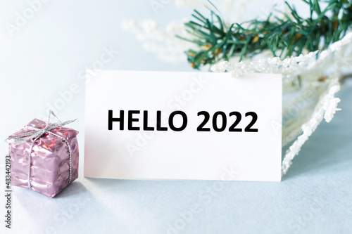 the text HELLO 2022 on the card next to the gift in the package and two spruce branches, festive background