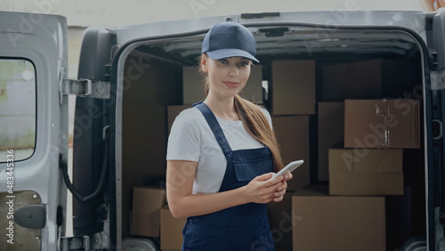 Delivery woman in overalls holding smartphone and looking at camera near cardboard boxes in car outdoors
