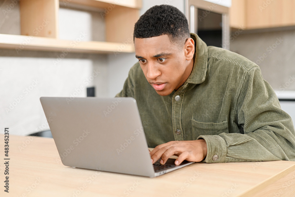 Serious focused determined young businessman or freelancer in casual clothes sitting at kitchen table, typing on laptop, working on project, researching, writing email to client Serious focused determined young businessman or freelancer in casual clothes sitting at kitchen table, typing on laptop, working on project, researching, writing email to client