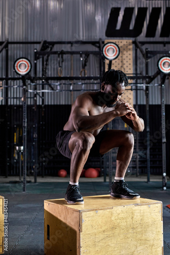 Fit shirtless african athletic man jumping up to plyometric wood box at gym, indoors, at modern gym. Handsome young afro american shirtless guy engaged in sport, doing exercises. side view