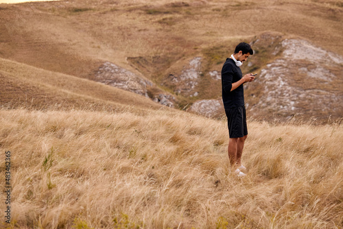 Athlete Caucasian Male In Shorts And T-shirt With Headphones Use Smartphone Before Jogging, Preparing To Train, Do Exercises, Turn On The Best Music For Workout. In Countryside