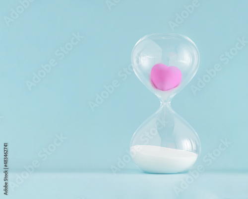2022. Minimal abstract creative concept with pink heart stuck in hourglass on isolated pastel blue background. Eternal or imperishable love idea. Gift or greeting card for wedding or Valentine’s Day.