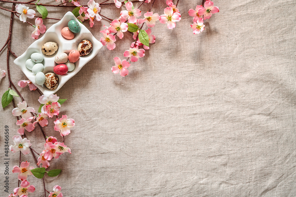 Fototapeta premium Easter background with colored easter eggs in egg stand and blooming branches on a beige background. Easter composition on a textured beige linen background.